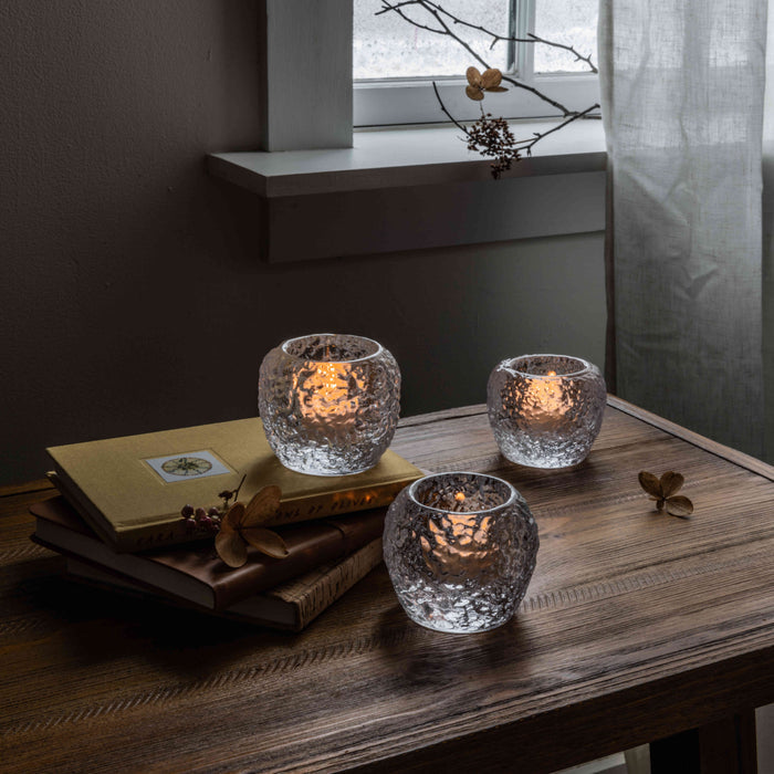 Three textured glass tealight holders with lit candles on a wooden table.