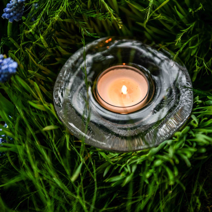 Lit tealight candle in gray metal holder surrounded by green foliage and small blue flowers.