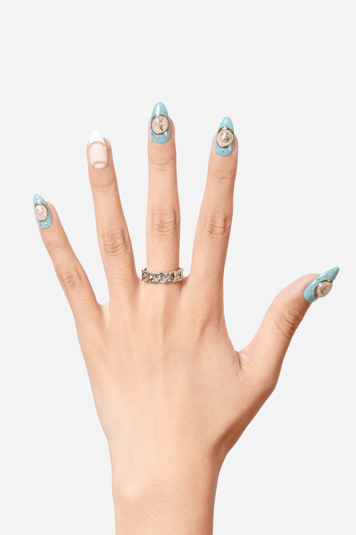 Hand showcasing "A Touch of Charm" press-on nails: light blue with gold charms & rhinestones, neutral accent nail. Silver chain ring.