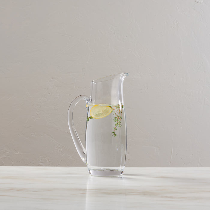 Clear glass pitcher with handle containing water, lemon slice, and fresh herbs.