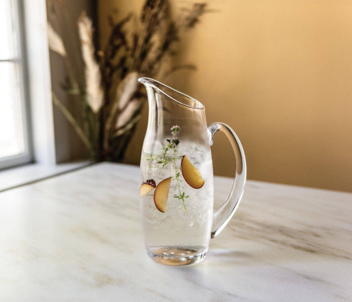 Clear glass pitcher filled with ice water, apple slices, and fresh herbs on marble surface.