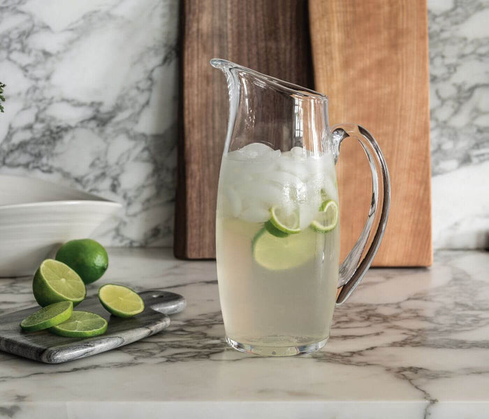 Clear glass pitcher filled with ice water and lime slices, positioned on marble surface.