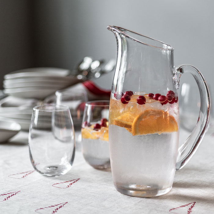 Clear glass pitcher filled with water, ice, orange slices, and cranberries, displayed with matching glasses.