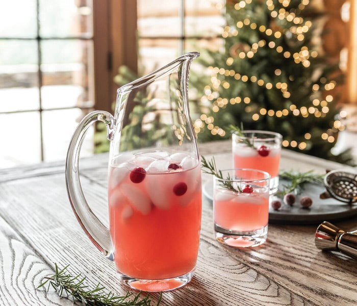 Clear glass pitcher filled with red beverage and cranberries, displayed with matching glasses on a wooden table.