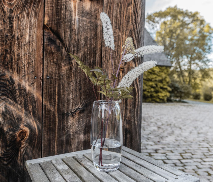 Clear glass cylindrical vase with white feathery stems and green foliage on wooden surface.