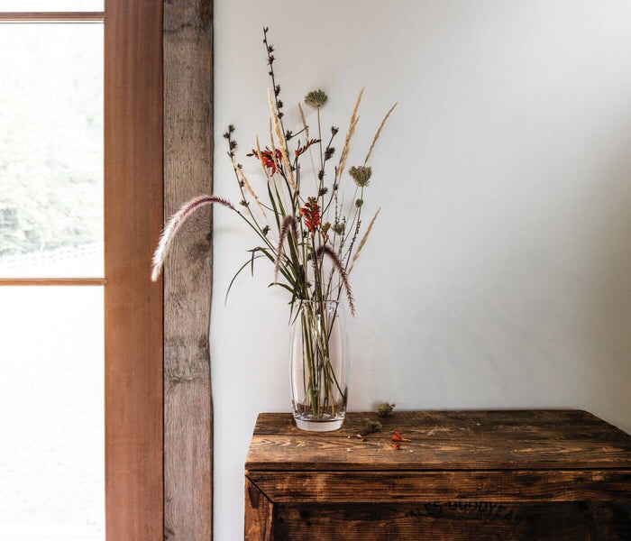 Clear glass cylinder vase filled with dried flowers and grasses on wooden surface.
