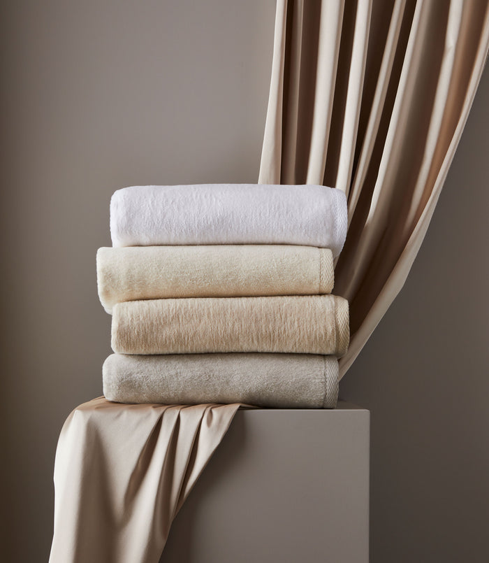 Stack of plush, 100% cotton All Seasons blankets in neutral shades with textured binding, displayed on a pedestal.