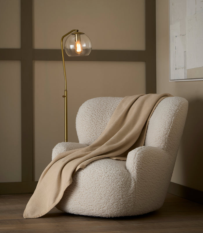 Plush beige 100% cotton blanket with textured binding draped over a white boucle armchair in a cozy room.