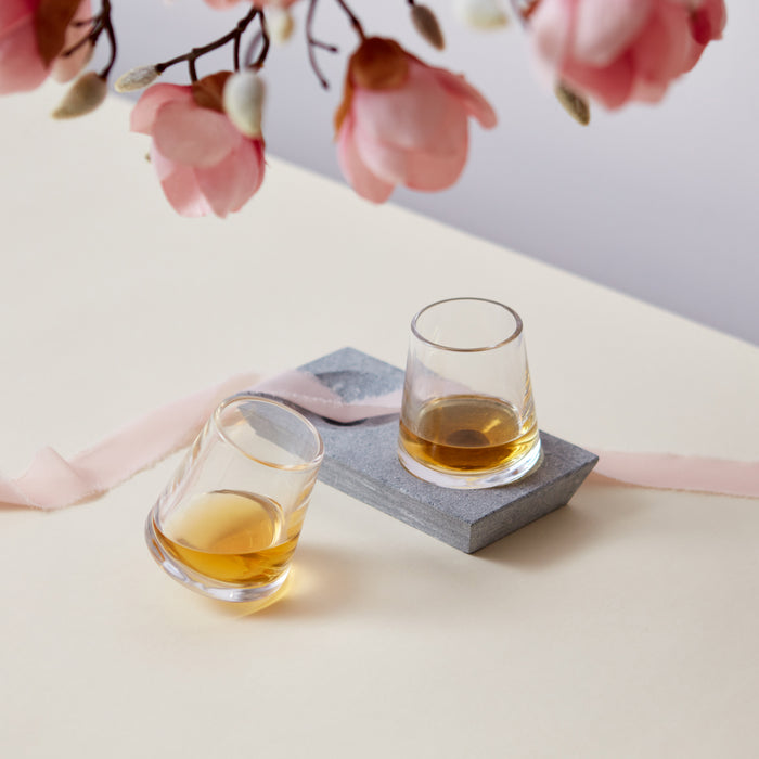Two whiskey glasses with amber liquid, one on a gray soapstone coaster, styled with pink flowers and ribbon.