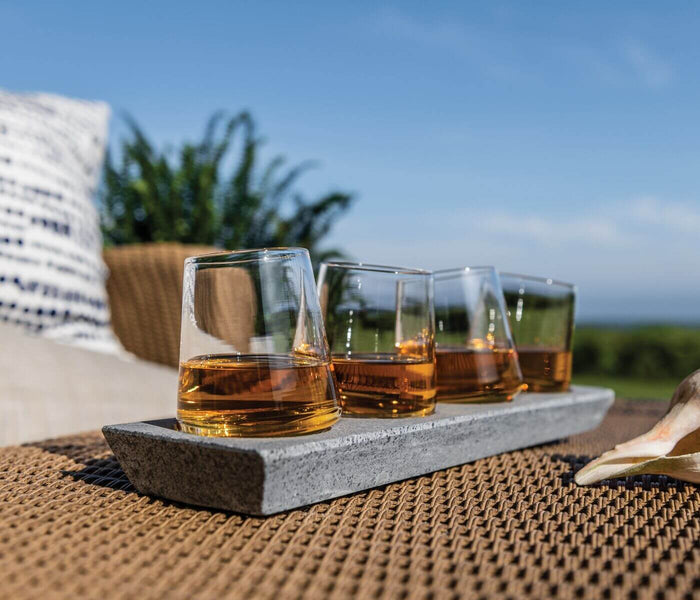 Four whiskey glasses with amber liquid sitting on a gray soapstone base outdoors.