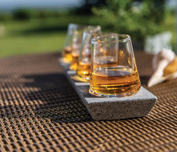 Four whiskey glasses filled with amber liquid, each seated on a gray soapstone base, arranged on a woven outdoor surface.