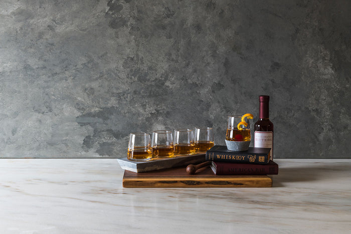 Four whiskey glasses with soapstone bases arranged on wooden holder with whiskey bottle and book.