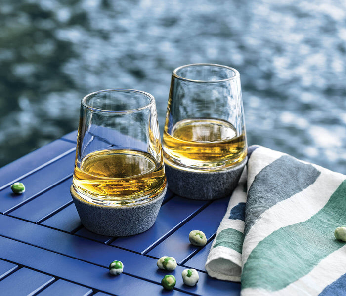 Two whiskey glasses with soapstone bases filled with amber liquid, set on blue surface with striped cloth and scattered berri