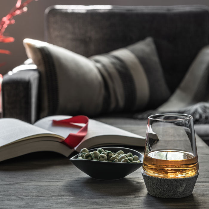Whiskey glass with soapstone base filled with amber liquid, beside an open book and bowl of nuts.