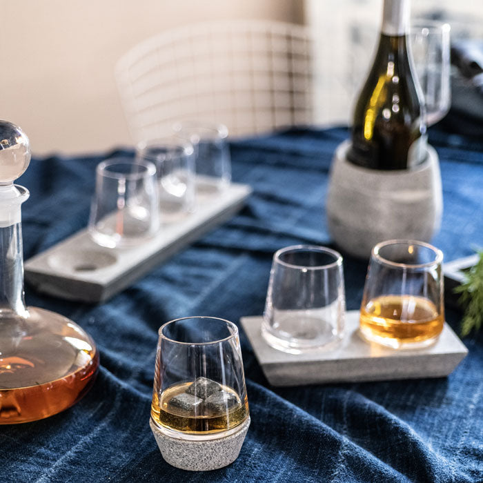 Whiskey glasses with soapstone bases arranged on a blue fabric, with a bottle in a soapstone holder and a decanter in the bac