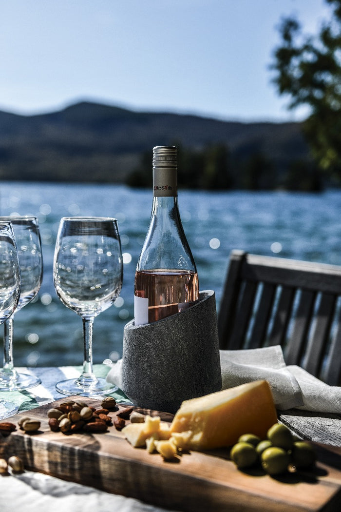 Gray soapstone wine chiller holding a rosé bottle, displayed on outdoor table with glasses, cheese, nuts, and mountain lake b