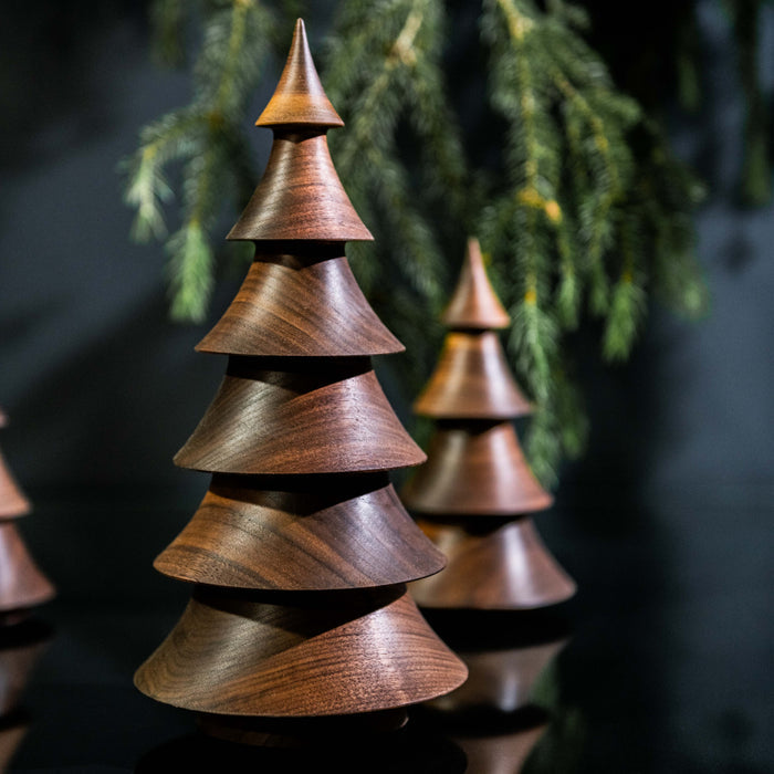 Stacked walnut wood tree sculpture with pointed top and evergreen branches in soft focus background.