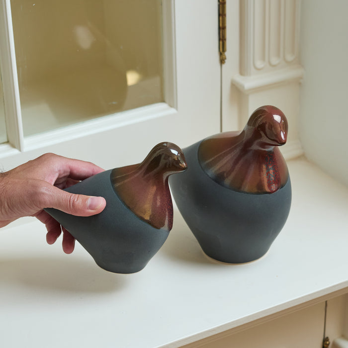 Two ceramic dove figurines with metallic bronze heads and matte gray bodies, one held in a hand.
