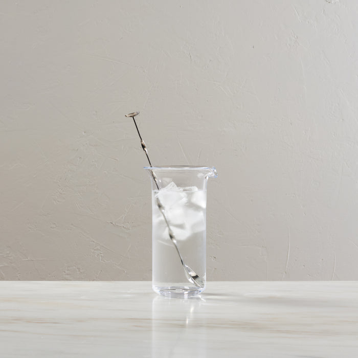 Clear glass tall pitcher containing ice water with a metal cocktail stirrer.