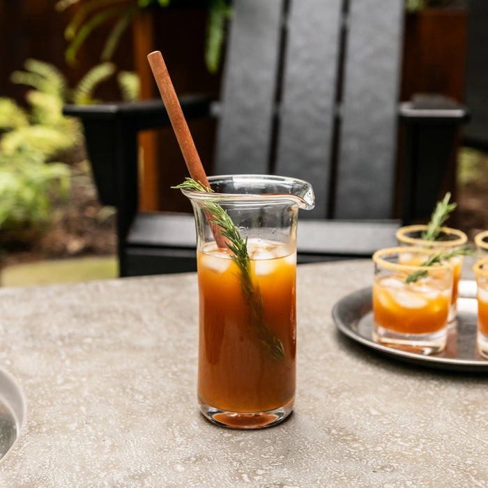 Clear glass pitcher filled with brown liquid, wooden stirrer, and fresh herb garnish on outdoor table.