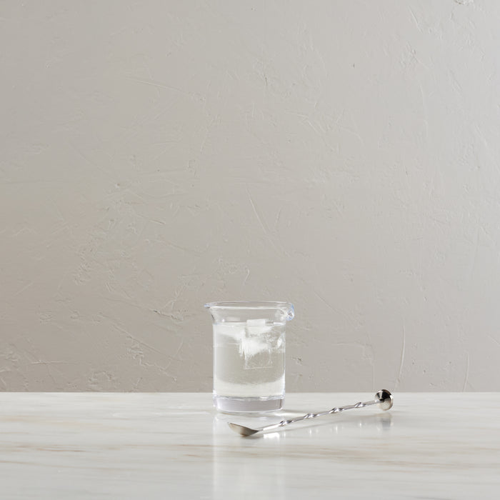 Clear glass pitcher with pouring spout, containing water and ice, displayed with a bar spoon.