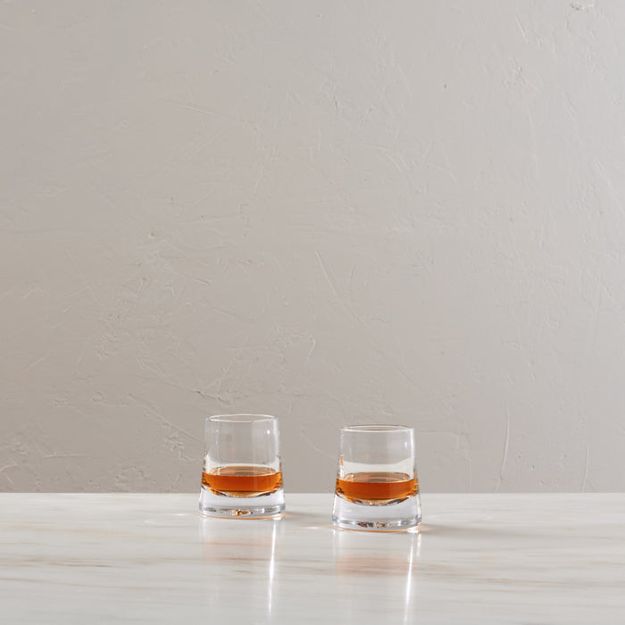 Two glasses filled with amber bourbon liquid on a white surface against a beige textured wall.