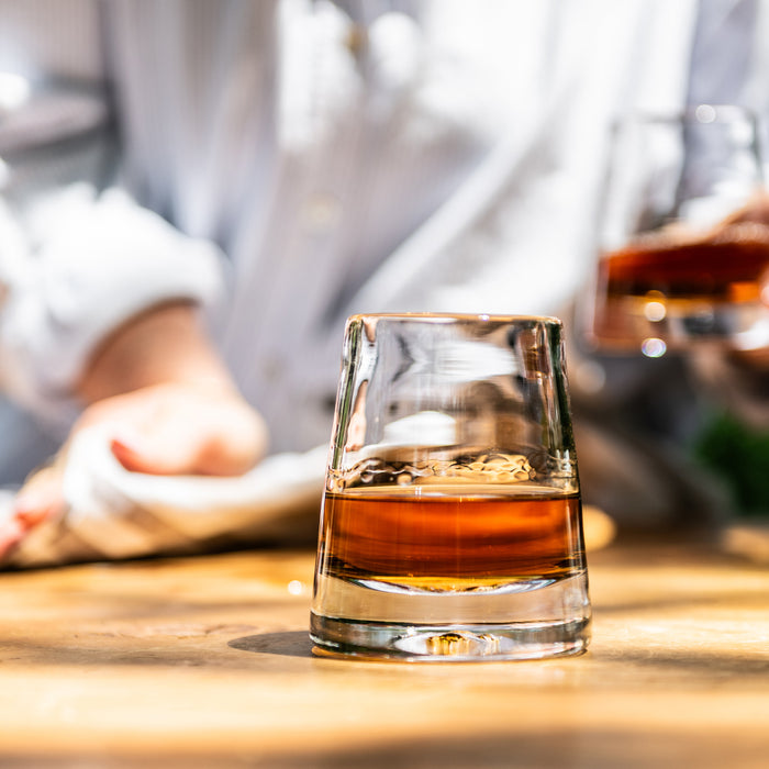 Glass of bourbon with ice on wooden surface, blurred bottles and person in background.
