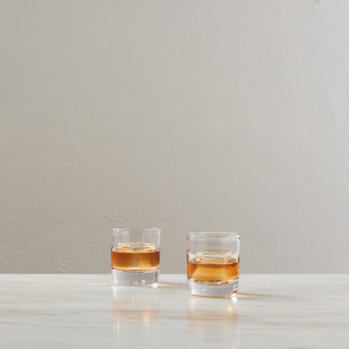 Two clear double old-fashioned glasses filled with amber liquid and ice cubes on a white surface.