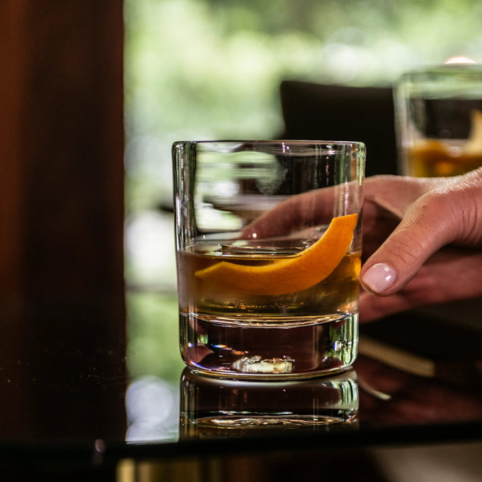 Double old-fashioned glass held in hand, filled with amber liquor and orange peel garnish.