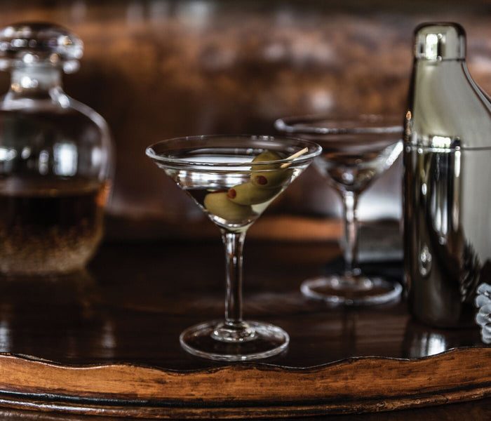 Two martini glasses with olives on a pick, cocktail shaker, and bottle on a wooden surface.