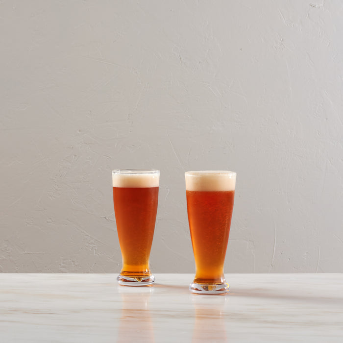 Two glasses of amber pilsner beer with cream-colored foam heads on a white surface.
