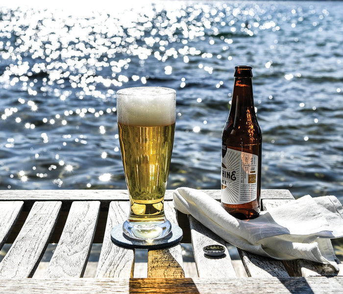 Pint glass of pilsner beer and brown bottle on whitewashed wooden dock with water and napkins in background.