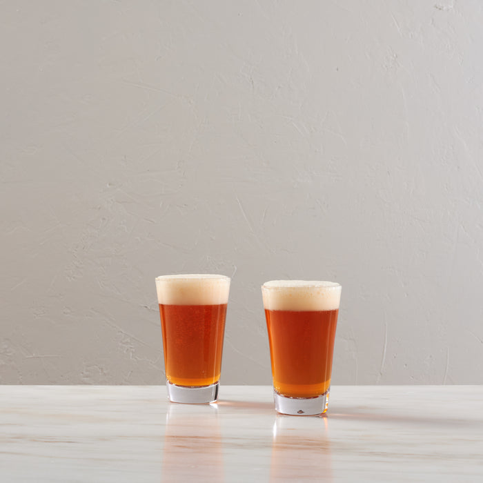 Two pint glasses filled with amber beer and topped with foam heads on a white surface.