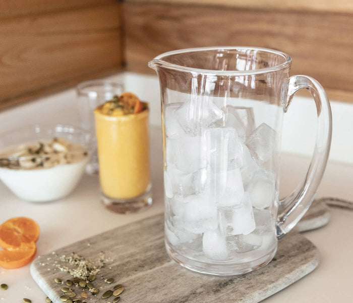 Clear glass pitcher filled with ice and water, with handle, on marble coaster.