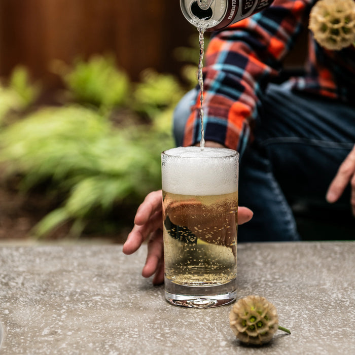 Beverage being poured into a clear glass tumbler with foam head, held by a hand outdoors.