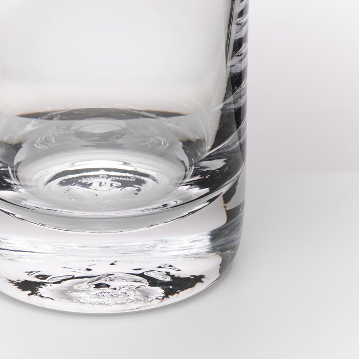 A close-up of a clear glass tumbler with a thick, rounded base and subtle etched or textured detailing on the side, shown against a clean white background.
