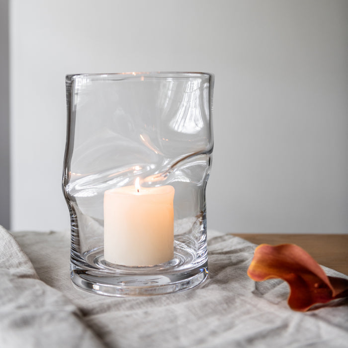 Clear glass hurricane with twisted design holding a lit cream-colored pillar candle on linen fabric.