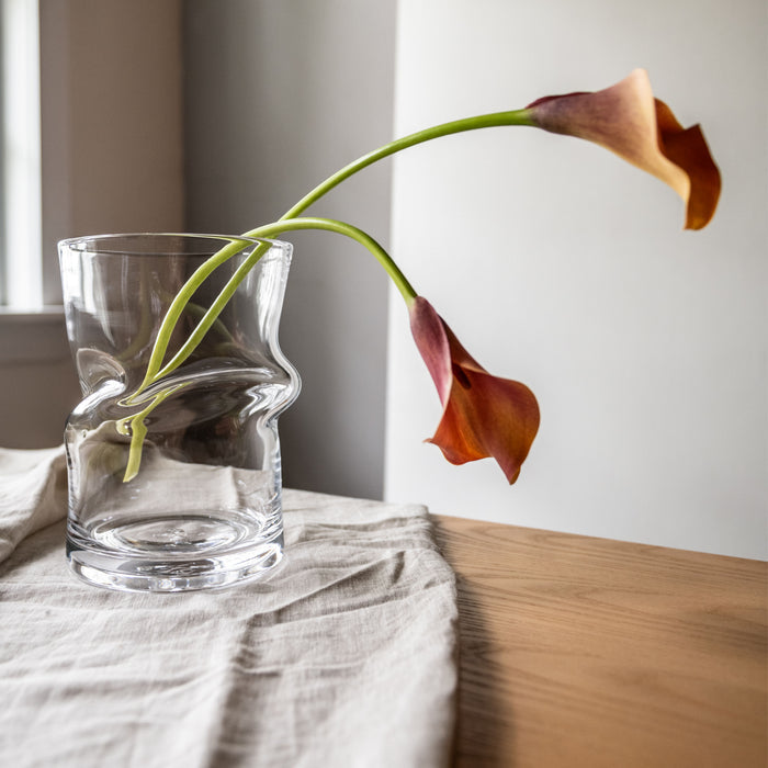 Clear glass hurricane vase with curved twisted shape holding two red and orange calla lilies with green stems.