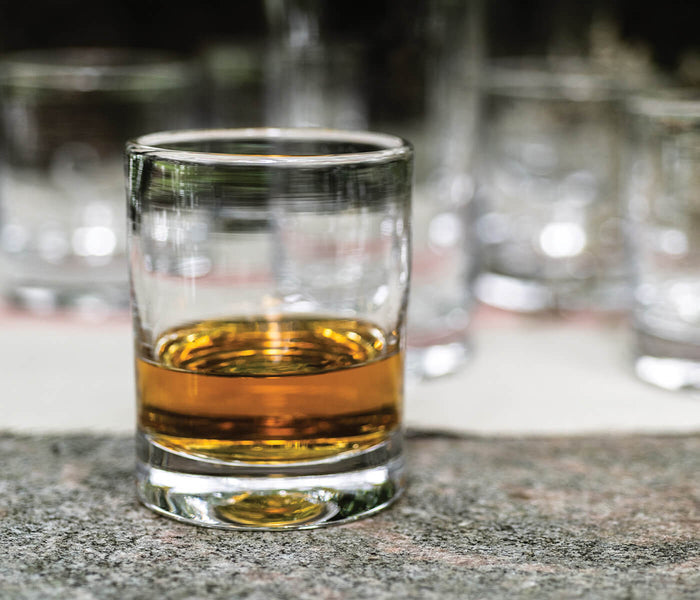 Two clear whiskey glasses with amber liquid, one in focus in foreground and others blurred in background.