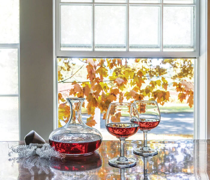 Glass wine decanter filled with red wine next to two wine glasses on a table by a window with autumn foliage visible outside.