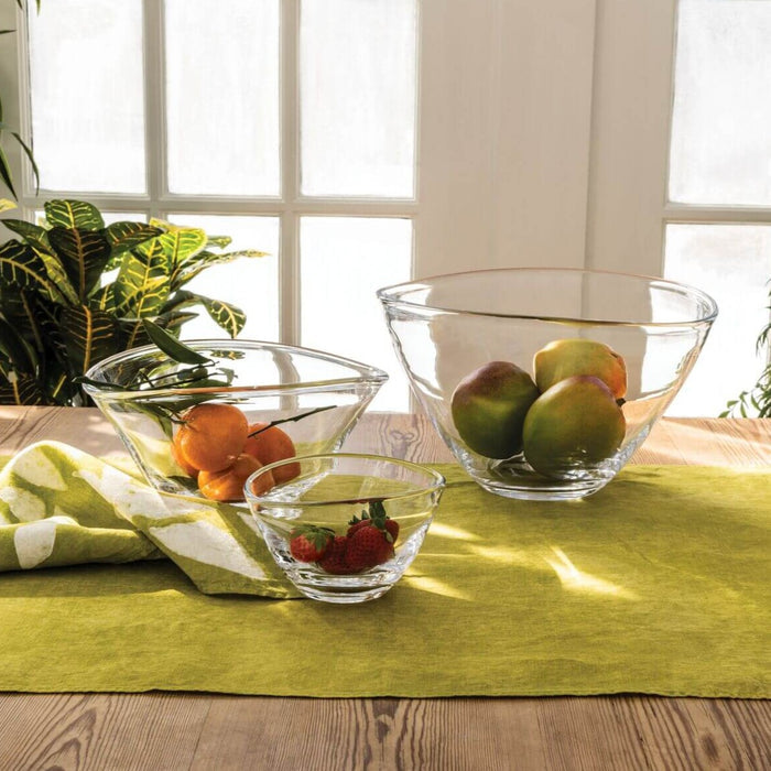 Three clear glass bowls in varying sizes filled with apples, oranges, and strawberries on a yellow table cloth.