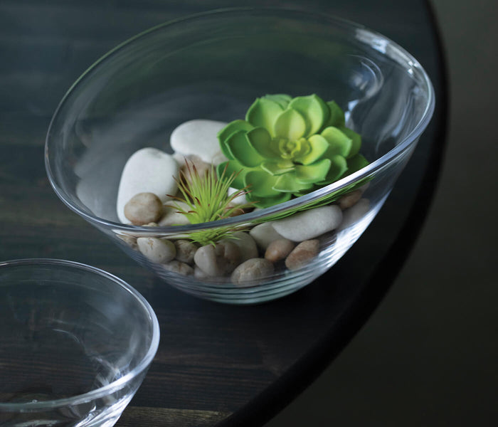 Clear glass bowl with curved sides containing green succulent, white stones, tan pebbles, and decorative grass.