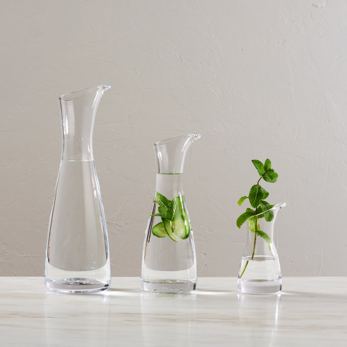 Clear glass carafe with tapered top and white band, shown in three sizes with water and fresh herbs inside.