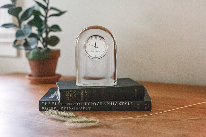 Clear glass arch-shaped clock with round dial displaying 1 o'clock, sitting on a dark book atop a wooden surface.