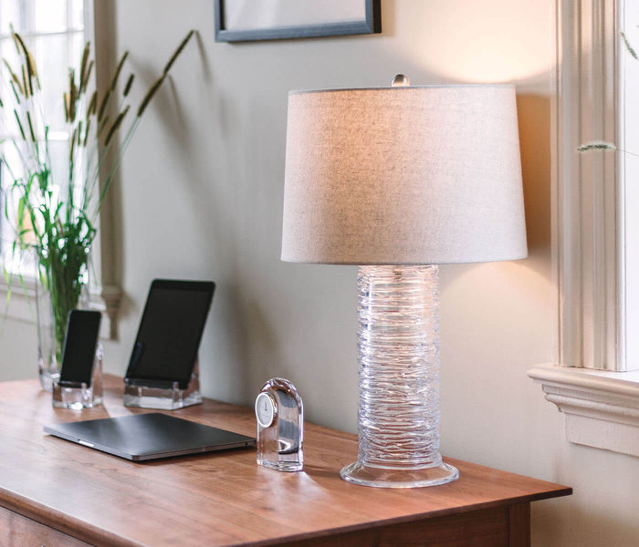 Clear cylindrical clock with textured glass base and cream linen drum shade on wooden desk.
