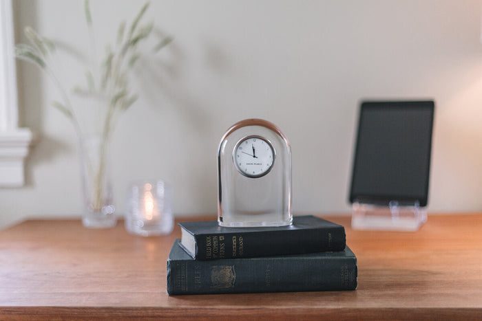 Clear glass arch-shaped clock with chrome frame and white dial, displayed on stacked black books.
