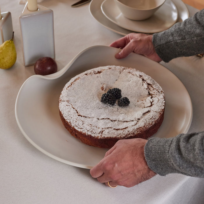 White wavy ceramic platter holding a dusted cake with blackberries, surrounded by fruit and tableware.