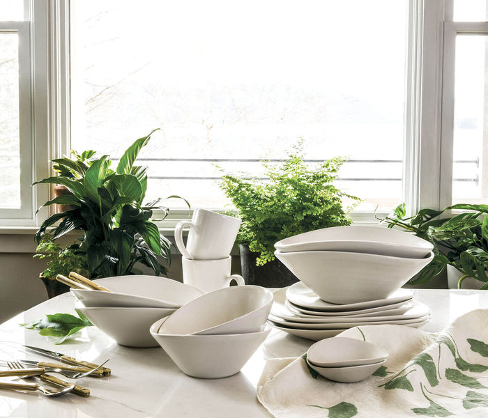 White ceramic serving bowl with curved sides displayed among matching tableware and plants on a table.
