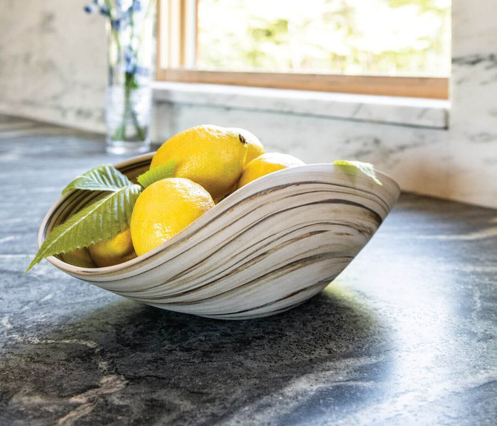 Wave-shaped ceramic bowl in cream and gray tones holding lemons and green leaves on a dark countertop.