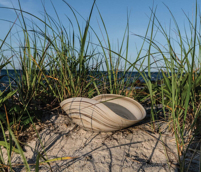 Cream-colored oval ceramic bowl resting on sand surrounded by beach grass.